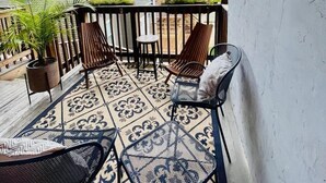 Een overdekt terras met een patroon vloer, twee rieten stoelen, een bijzettafeltje en een potplant.