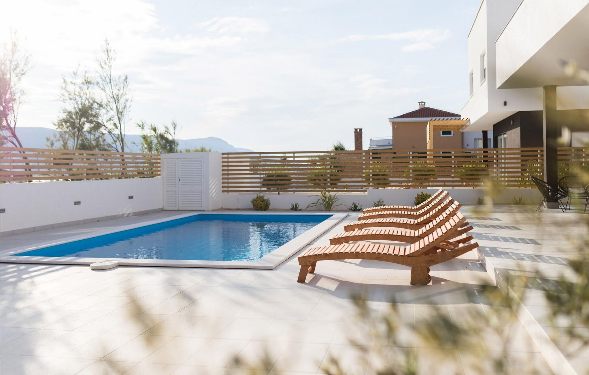 A modern outdoor pool area with a wooden bench, a white fence, and a building in the background.