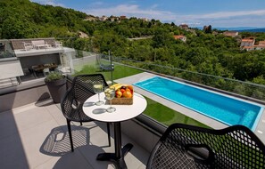 Eine Dachterrasse mit Pool, zwei Stühlen, einem Tisch mit Obst und Getränken und Blick auf eine Hügelkuppe mit Häusern.