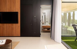 A modern living room with a flat-screen TV mounted on a wooden wall, a dark door leading to a bathroom, and a glass door to a balcony with a view of a lawn and trees. - Amazing home in Zadar with sauna (Zadar)