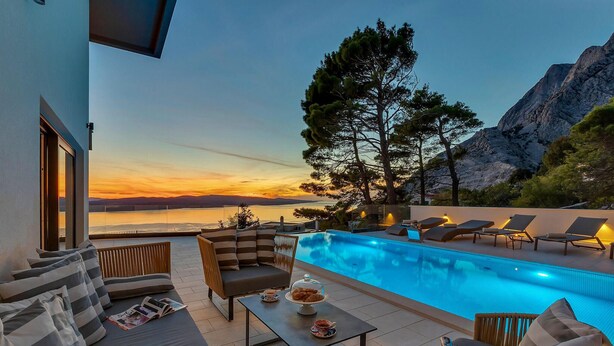 A poolside area with lounge chairs, a table with a cake and cups, and a view of a sunset over a body of water with mountains in the background.