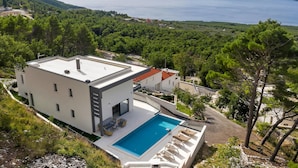 A modern two-story house with a swimming pool, surrounded by lush greenery and overlooking a large body of water.