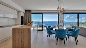 A modern kitchen with a wooden island, a dining area with blue chairs, and a large window offering a view of the sea and mountains.