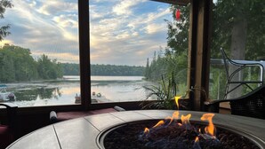 Un foyer avec des flammes, une vue sur un lac et un fauteuil près de la fenêtre.