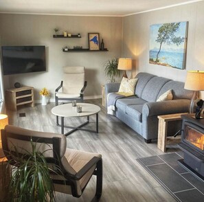 Ein modernes Wohnzimmer mit Sofa, Sessel und kleinem Tisch. Ein Kamin, ein an der Wand befestigter Fernseher und ein Gemälde sind ebenfalls vorhanden.