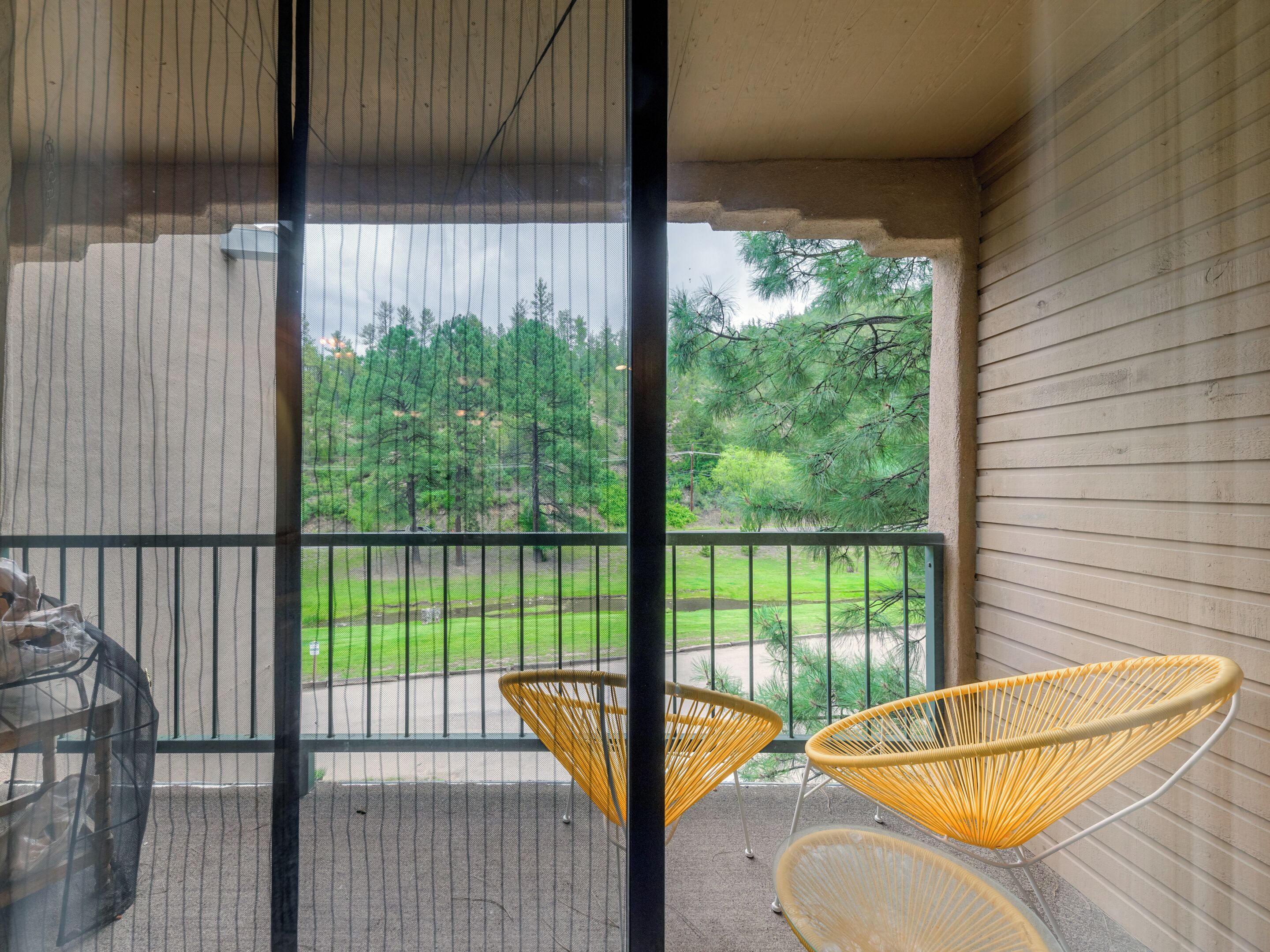 A balcony with a view of a grassy area and trees. There are two wicker chairs and a glass table.