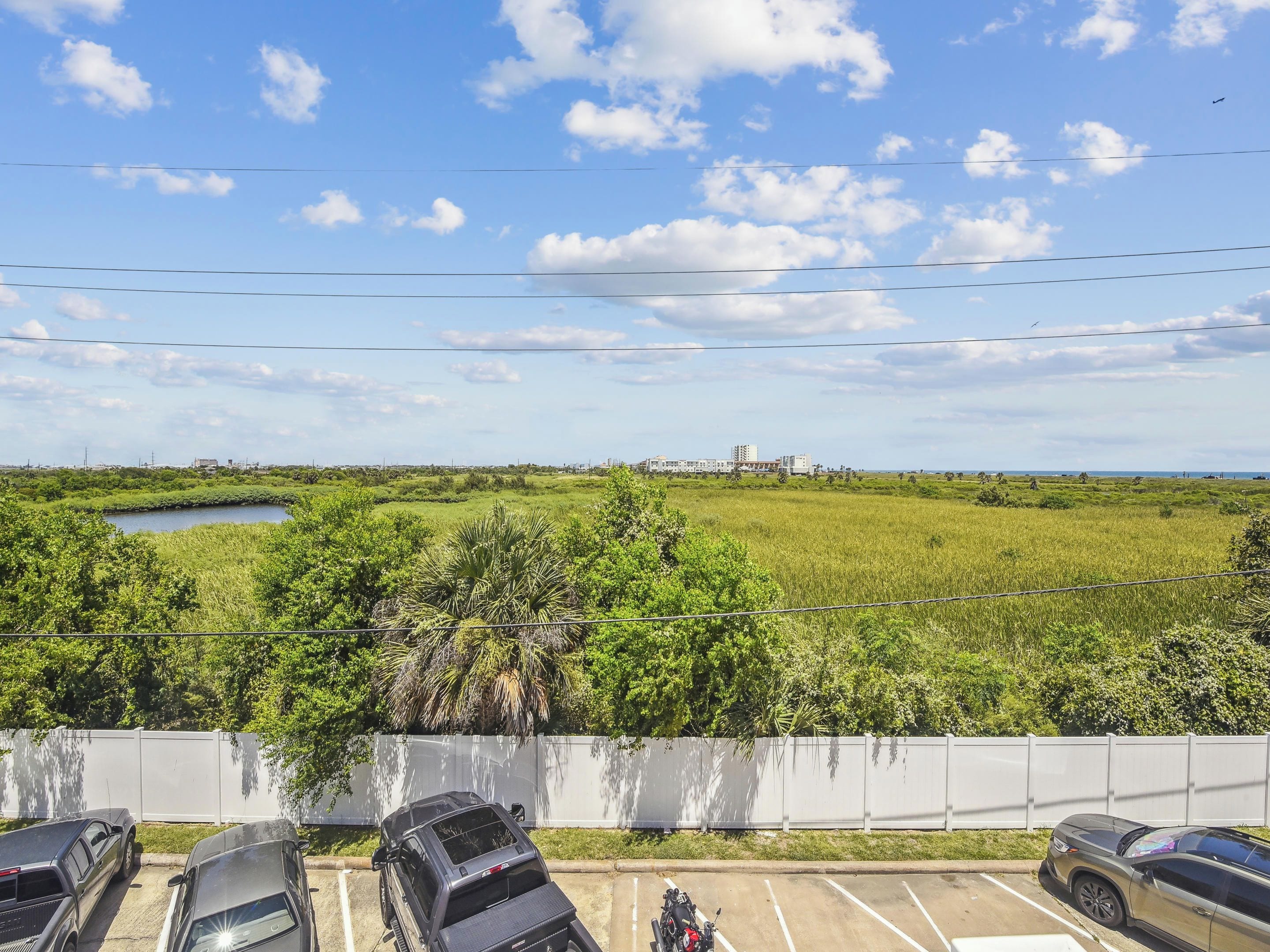 A parking area with several cars and a motorcycle, a grassy field with a small body of water, and a distant view of buildings and a tall structure.