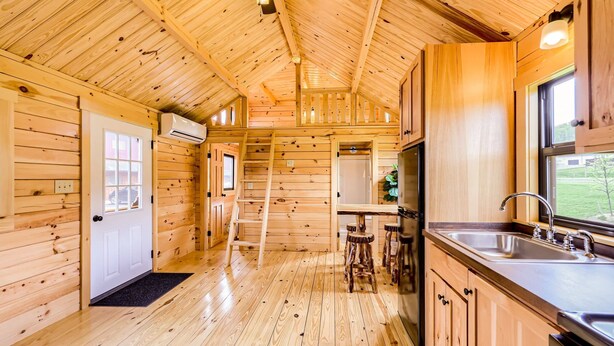 A wooden cabin interior with a kitchen area featuring a sink, stools, and a ladder leading to a loft.