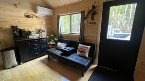 A cozy cabin interior with a wooden ceiling, a black sofa with cushions, a coffee station, and a window with a view of the forest.