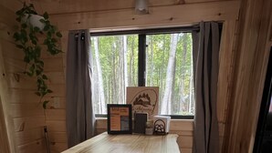 A wooden cabin interior with a large table, stools, a window with curtains, and a welcome sign.