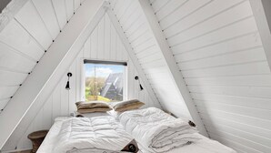 A bedroom with a sloped ceiling, a bed with white bedding, two wall-mounted lamps, and a window with a view of the outside.