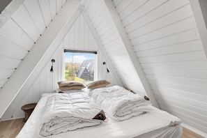 Ein Schlafzimmer mit schrägen Wänden, einem Bett mit weißer Bettwäsche, zwei Wandlampen und einem Fenster mit Blick nach draußen.