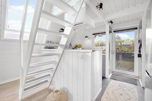 Un intérieur moderne avec un escalier en bois blanc, un coin cuisine et une porte vitrée donnant sur une terrasse extérieure.
