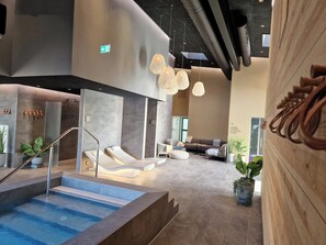 Ein moderner Innenpoolbereich mit Liegefläche, Sitzgelegenheiten und einer Treppe zu einem Spa-Bereich.