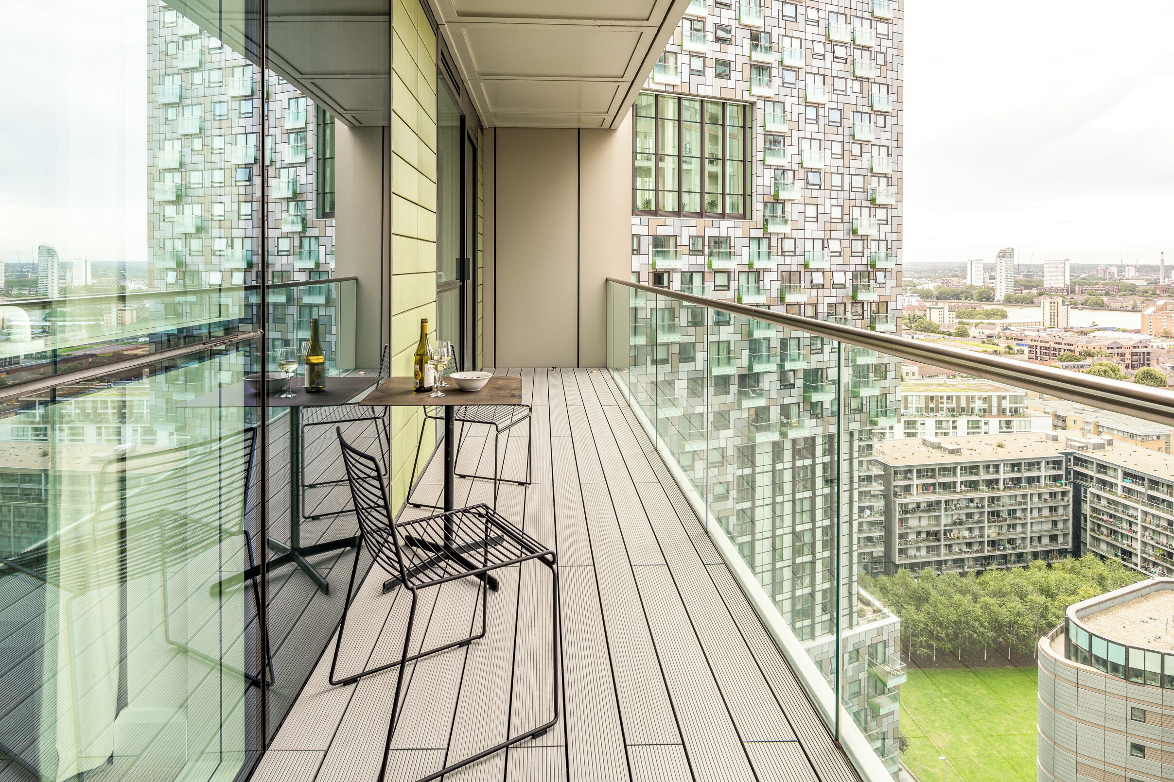 A modern balcony with a glass railing, a small table with a bottle and glasses, and a chair.
