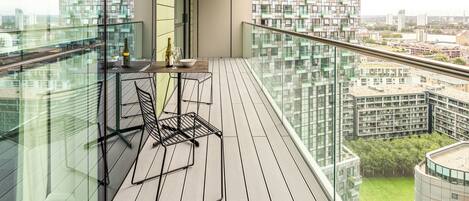 A modern balcony with a glass railing, a small table with a bottle and glasses, and a chair.