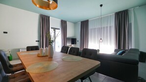 Ett modernt vardagsrum med ett matbord i trä, bekväma sittplatser, en platt-TV och stora fönster med gardiner.