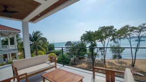 Ein Balkon mit Holzmöbeln, Blick auf den Strand und eine reetgedeckte Struktur in der Ferne.