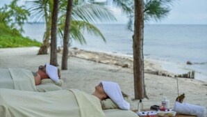 Eine Strandansicht mit zwei Personen, die auf Handtüchern liegen, einem Tisch mit Gegenständen und Palmen im Hintergrund.