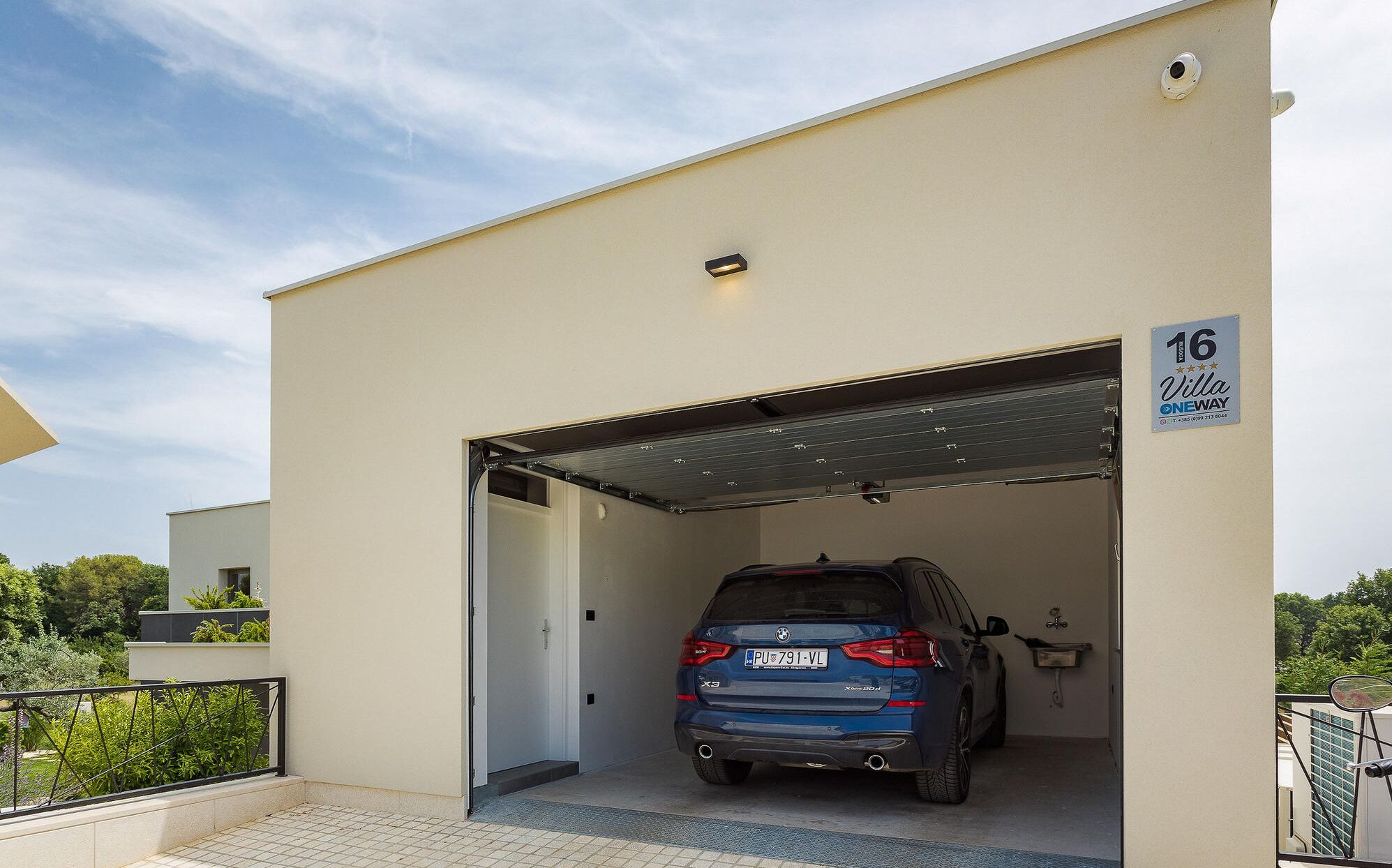 En moderne bygning med garageindgang, en blå bil parkeret indeni, og et skilt der angiver, at bygningen er en '16 Villa OneWay'.