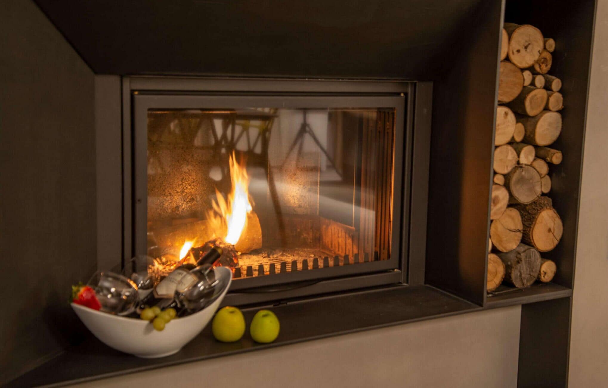 A fireplace with logs, a bowl of fruit, and a glass of wine on a mantel.