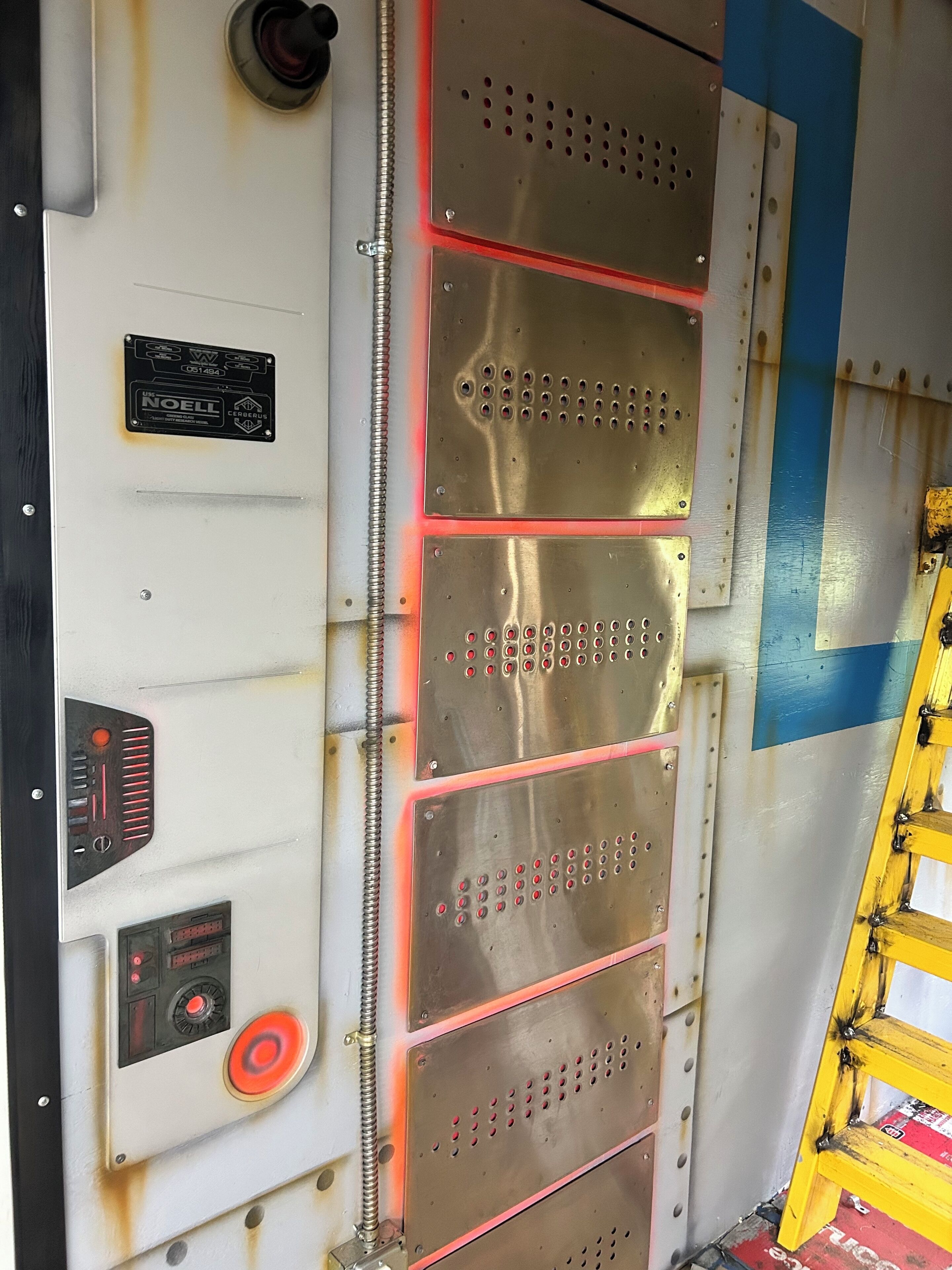 A metallic elevator interior with a control panel, emergency stop button, and a yellow safety railing.
