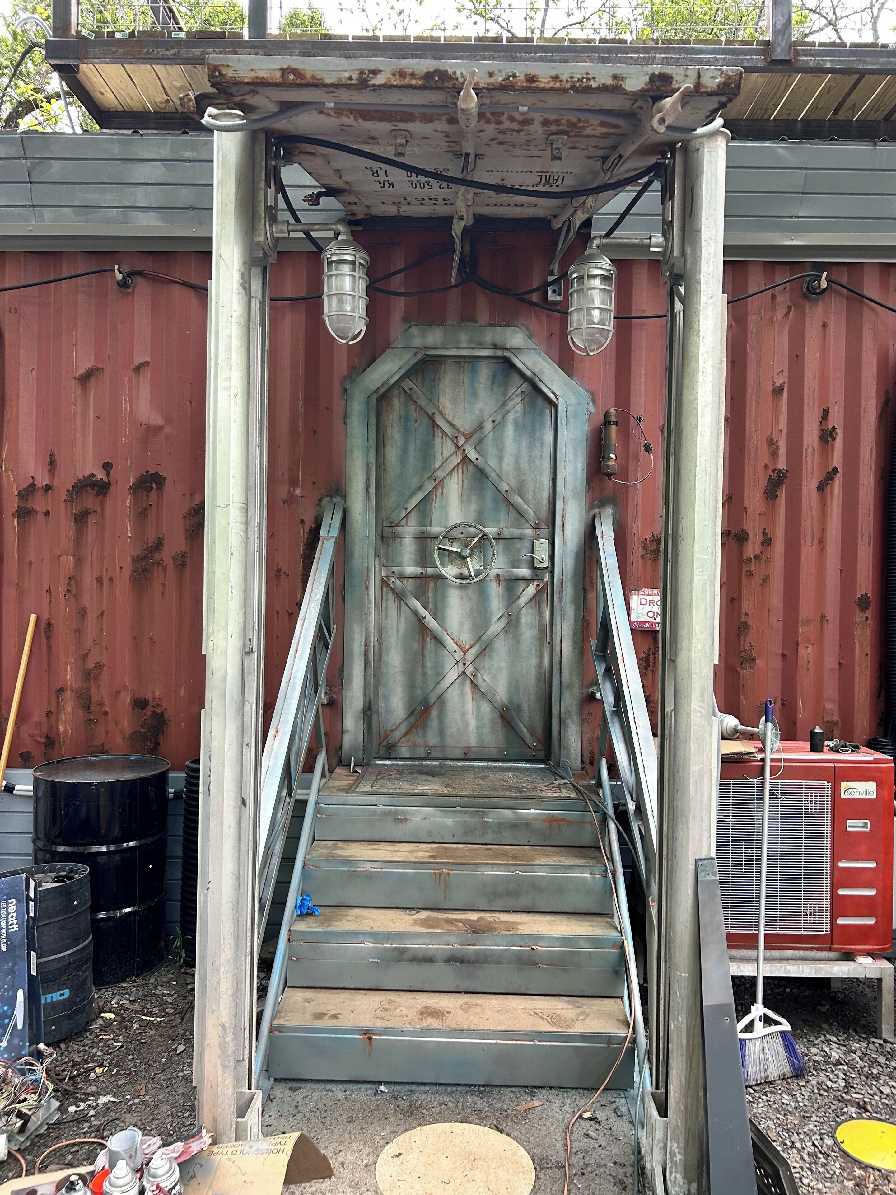 Un escalier en métal mène à une grande porte rouillée, dont le motif est constitué de traverses en forme de croix.