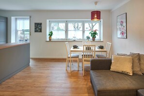 A modern living room with a sofa, a dining table with chairs, a kitchen area, and a large window with plants.