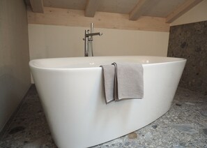 Una grande vasca da bagno freestanding in un bagno con pavimento in pietra e travi in legno al soffitto.
