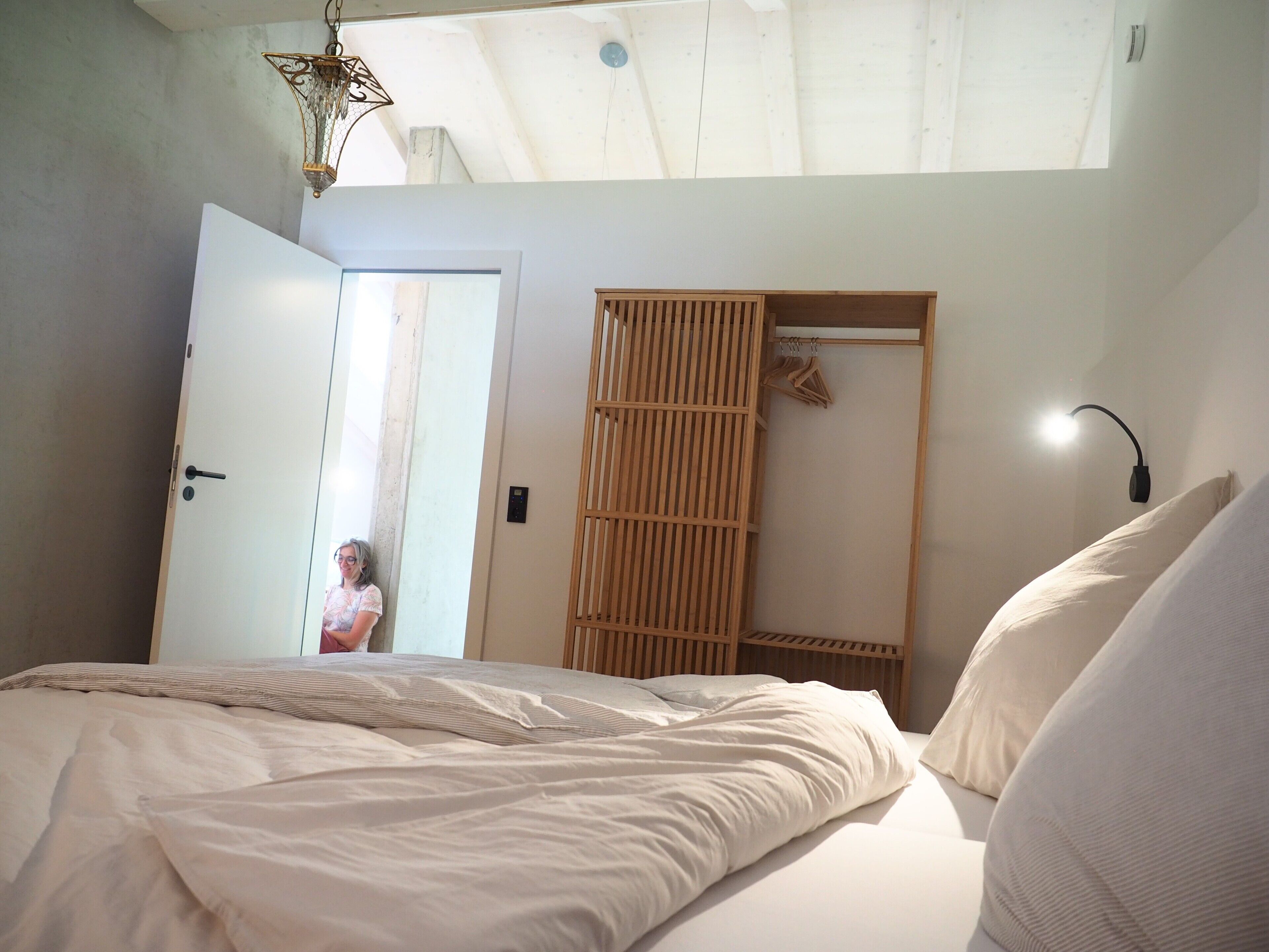 A bedroom with a bed, a wooden wardrobe, a hanging light fixture, and a door leading to another room.