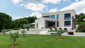 Una casa moderna de dos pisos con techo plano, ventanas amplias y un césped cuidado con arbolitos.