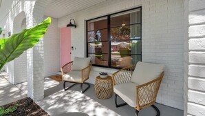 Eine Veranda mit zwei Korbstühlen und einem kleinen runden Tisch, weißen Ziegelwänden und einer großen Grünpflanze.