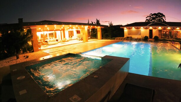 A swimming pool with a hot tub area, a covered patio with seating, and a building with warm lighting. - Llano Grande (Rionegro)