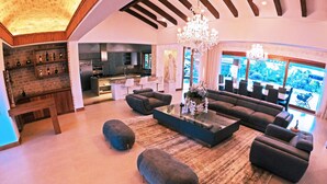 A spacious living room with a large sectional sofa, ottomans, and a coffee table. There is a chandelier, a fireplace, and a kitchen visible in the background. - Llano Grande (Rionegro)