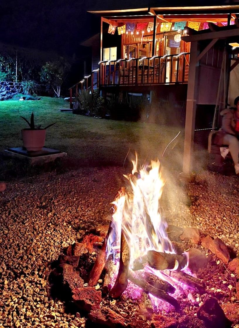 Un hogar con leña, rodeado de grava, frente a un edificio con terraza y luces colgantes.