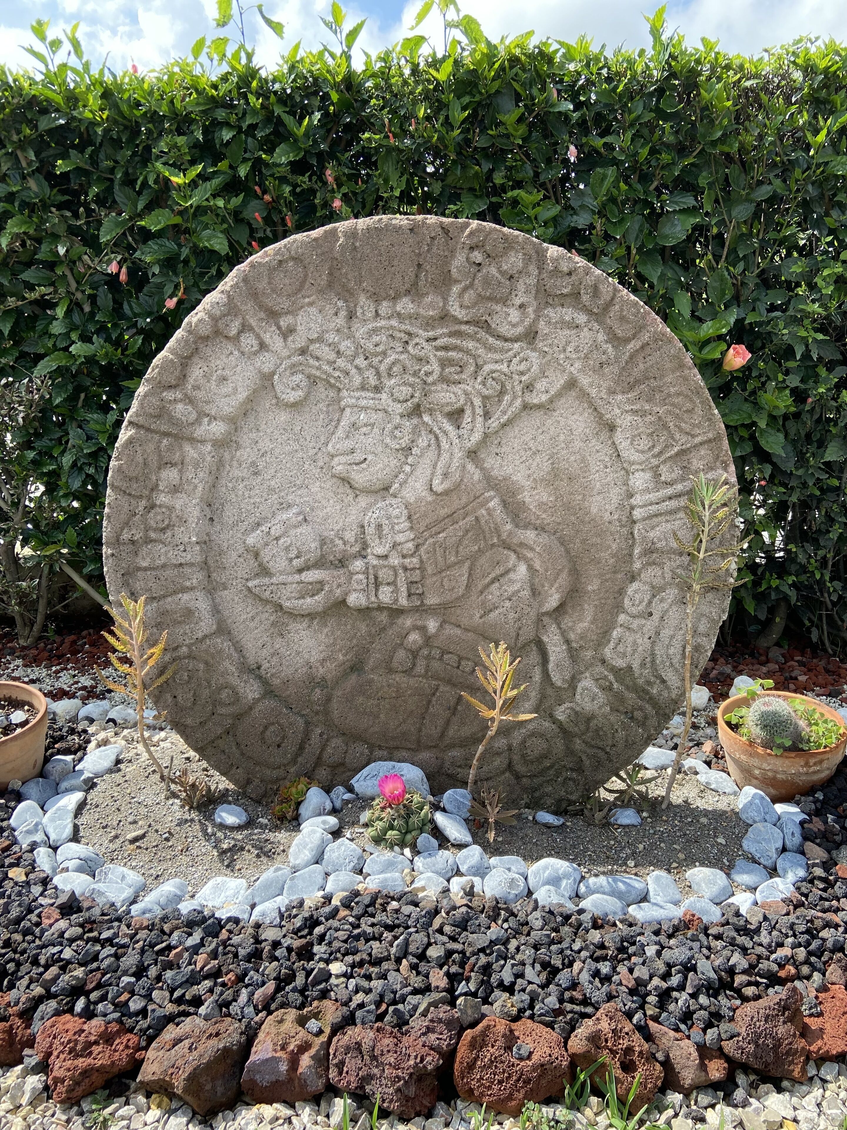 Uma grande escultura de pedra com detalhes complexos, cercada por um jardim com diversas plantas e rochas.
