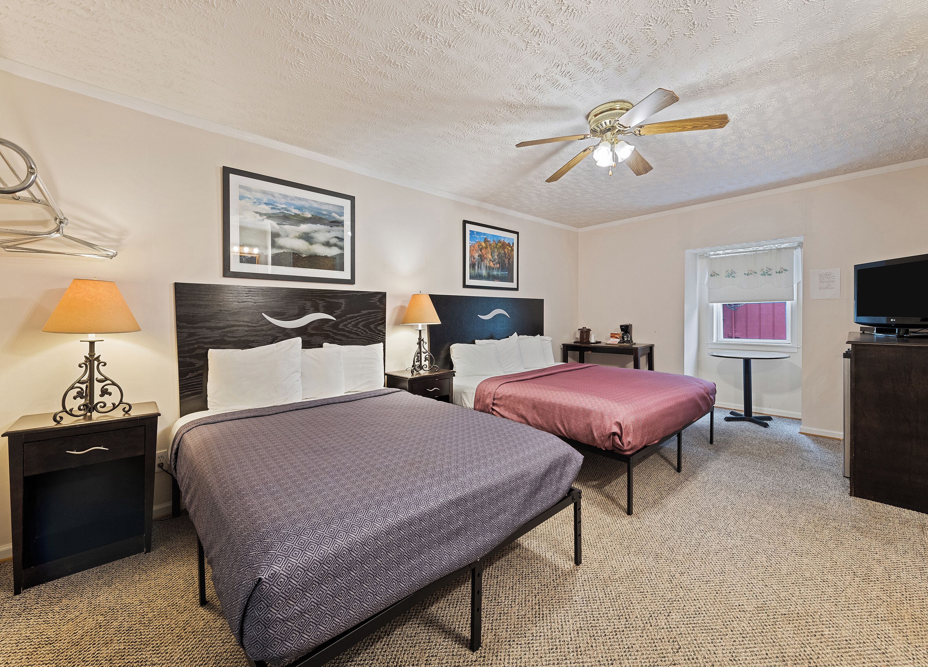 Une chambre d’hôtel avec deux lits, un ventilateur de plafond, une télévision et une petite table avec une lampe.