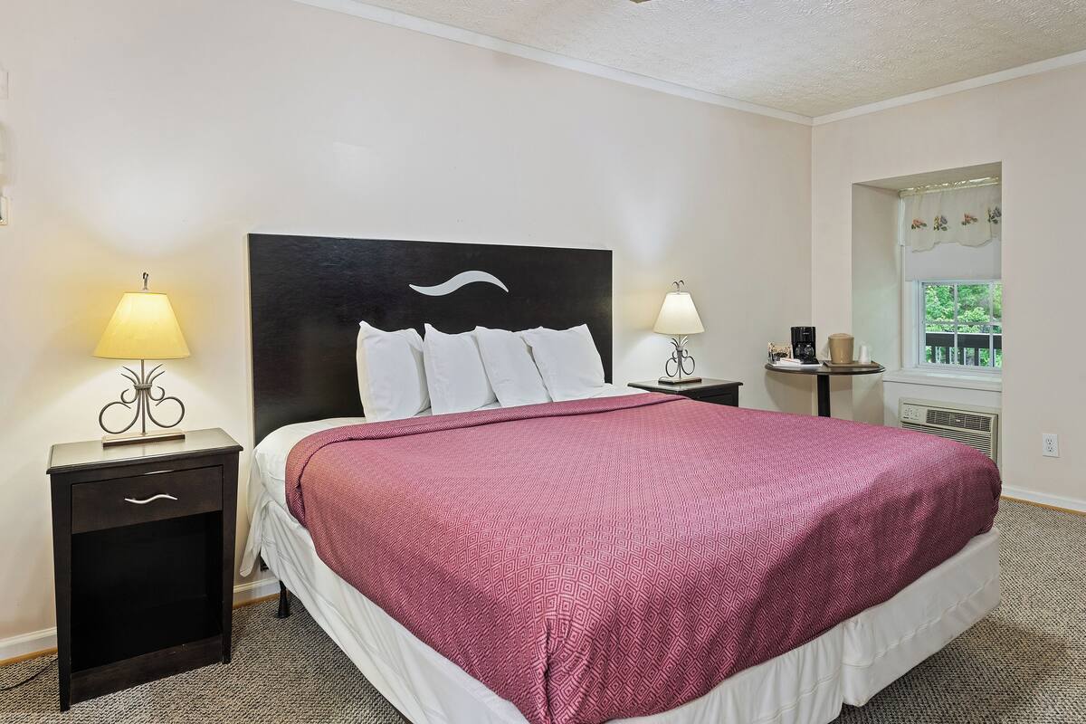 A hotel room with a large bed, two bedside tables with lamps, a ceiling fan, a window with a view, and a small table with a coffee maker.