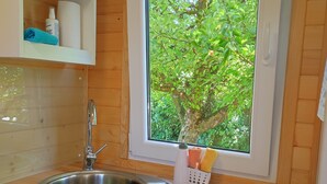 Une cuisine dotée d’un évier en acier inoxydable, de meubles en bois, d’une fenêtre donnant sur un espace verdoyant, et d’un plan de travail sur lequel se trouvent une planche à découper et un couteau.