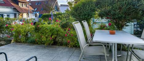 En terrasse med bord og stole, omgivet af grønt og udsigt til bygninger.