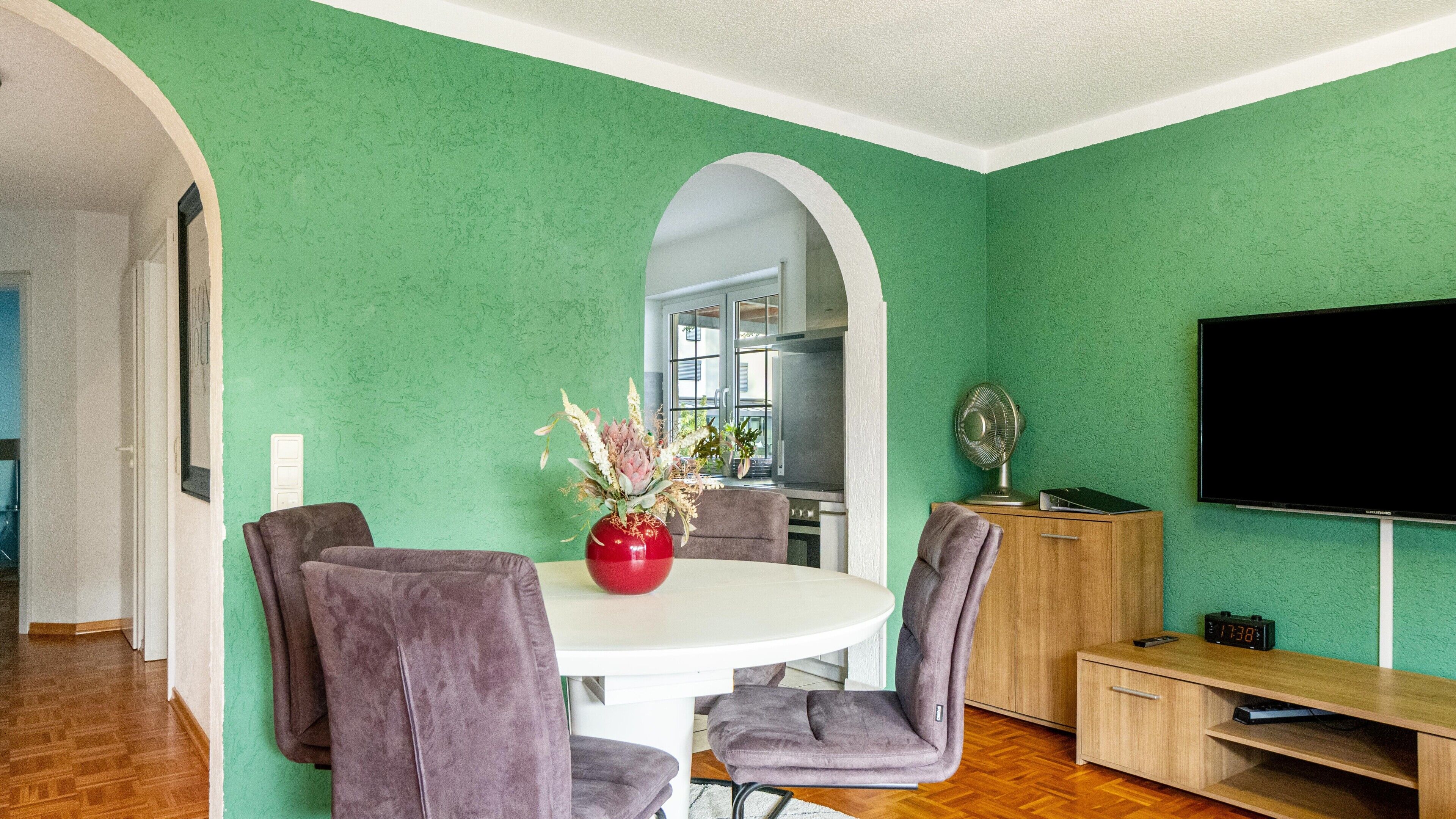 Ein Essbereich mit einem weißen Tisch, violetten Stühlen, einer roten Vase mit Blumen, einer Küche im Hintergrund und einem an der Wand montierten Fernseher.