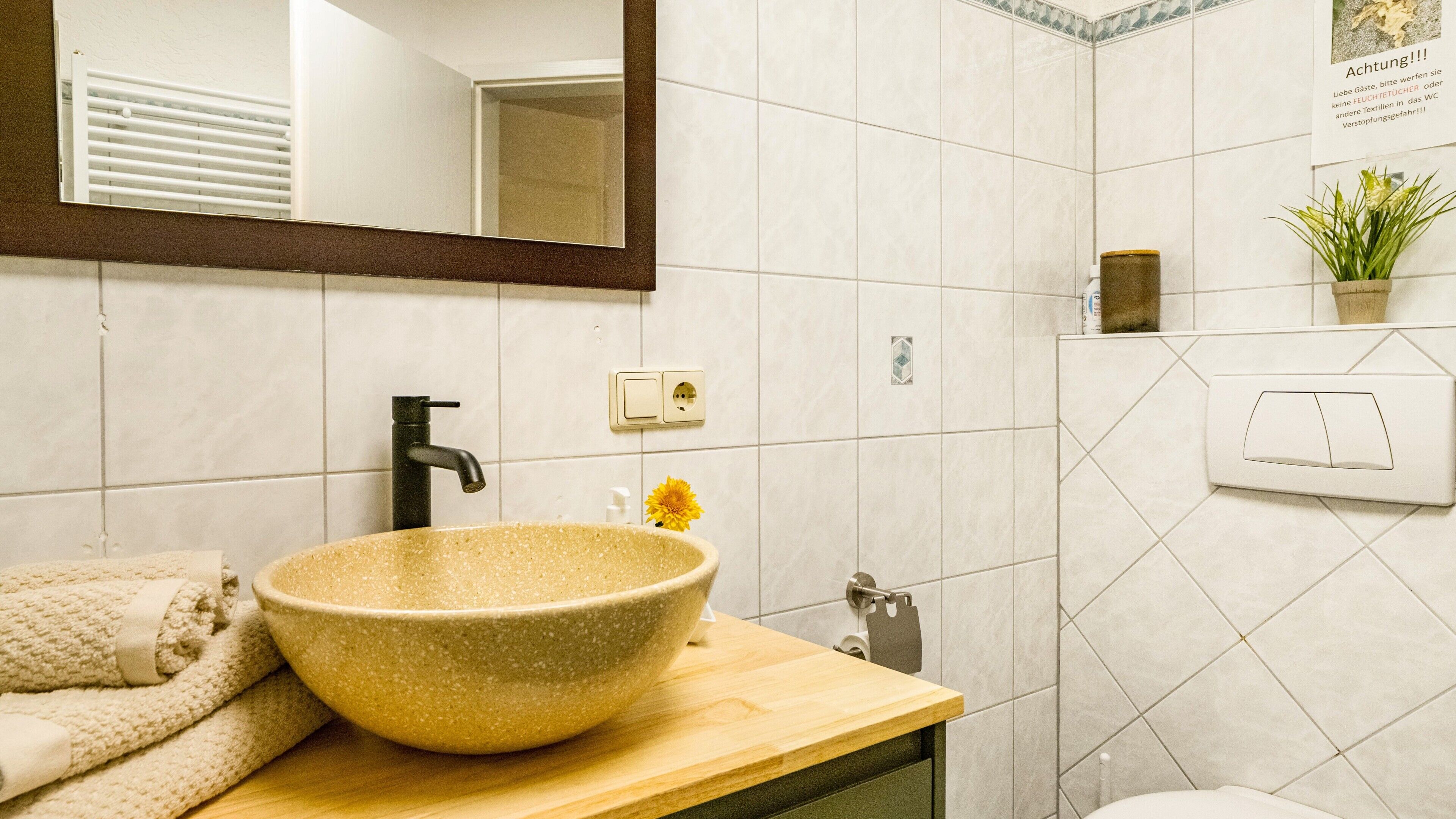 A bathroom with a beige sink, a black faucet, a towel, a flower, and a mirror.