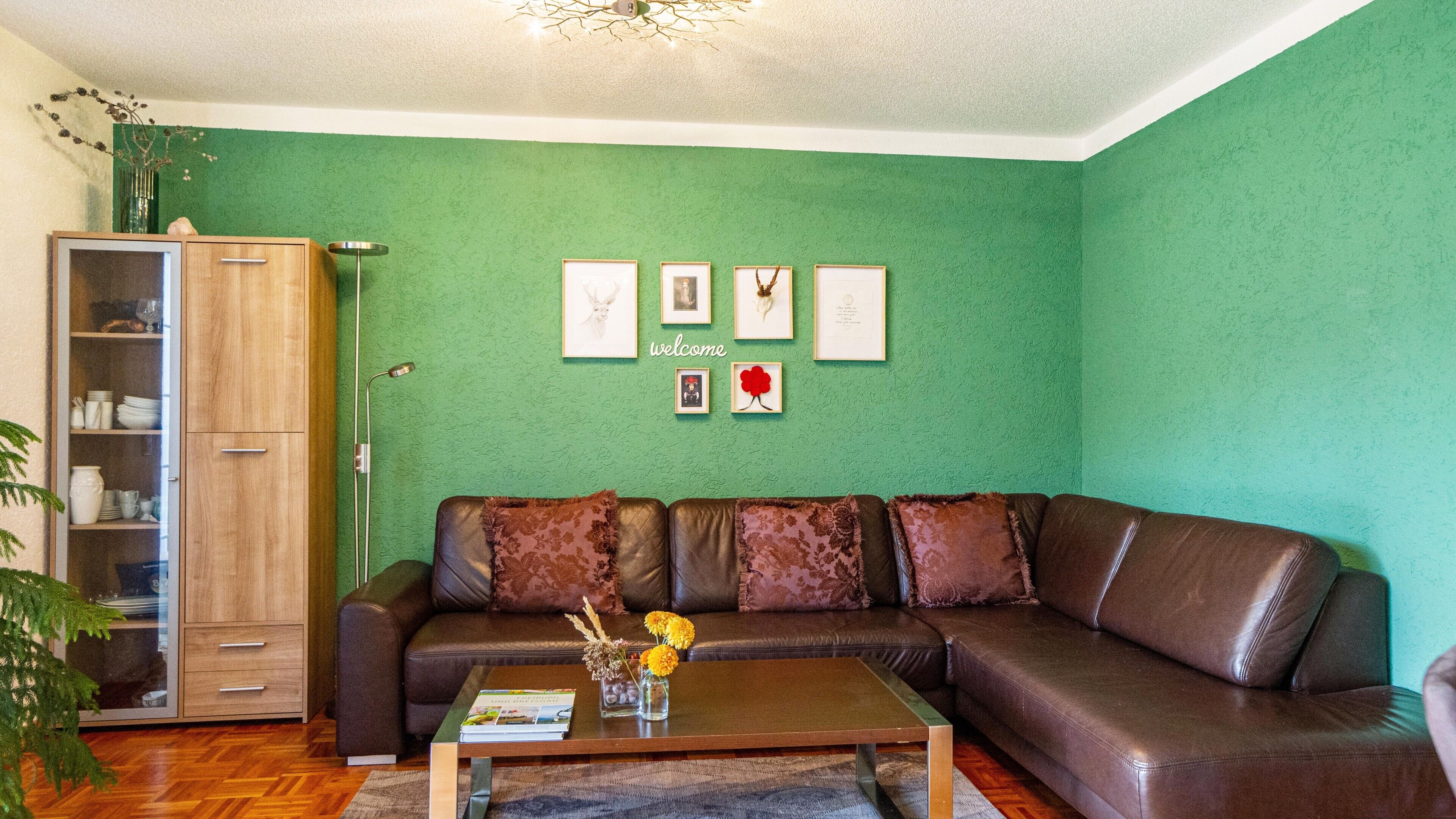 A living room with a green wall, a brown leather sofa, a wooden cabinet, and a coffee table with a book and flowers.