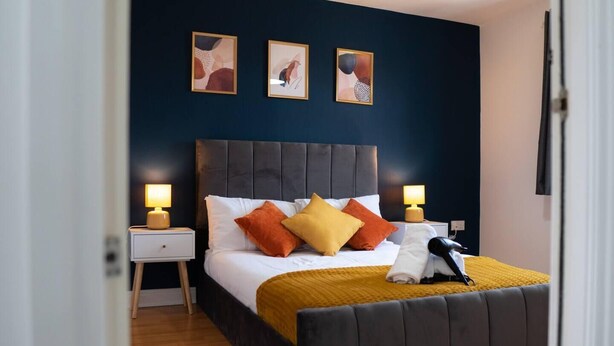 A modern bedroom with a dark headboard, a bed with white linens and multiple pillows, two bedside tables with lamps, and three framed artworks on the wall.