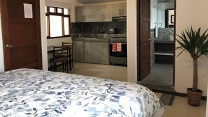 Une chambre avec un lit, une petite table et une chaise. On y trouve également un coin cuisine avec des placards et une cuisinière. Une plante en pot est visible.