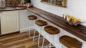 Een keuken met een rustieke bakstenen muur, een houten aanrechtblad, een grote spiegel en drie barstoelen.