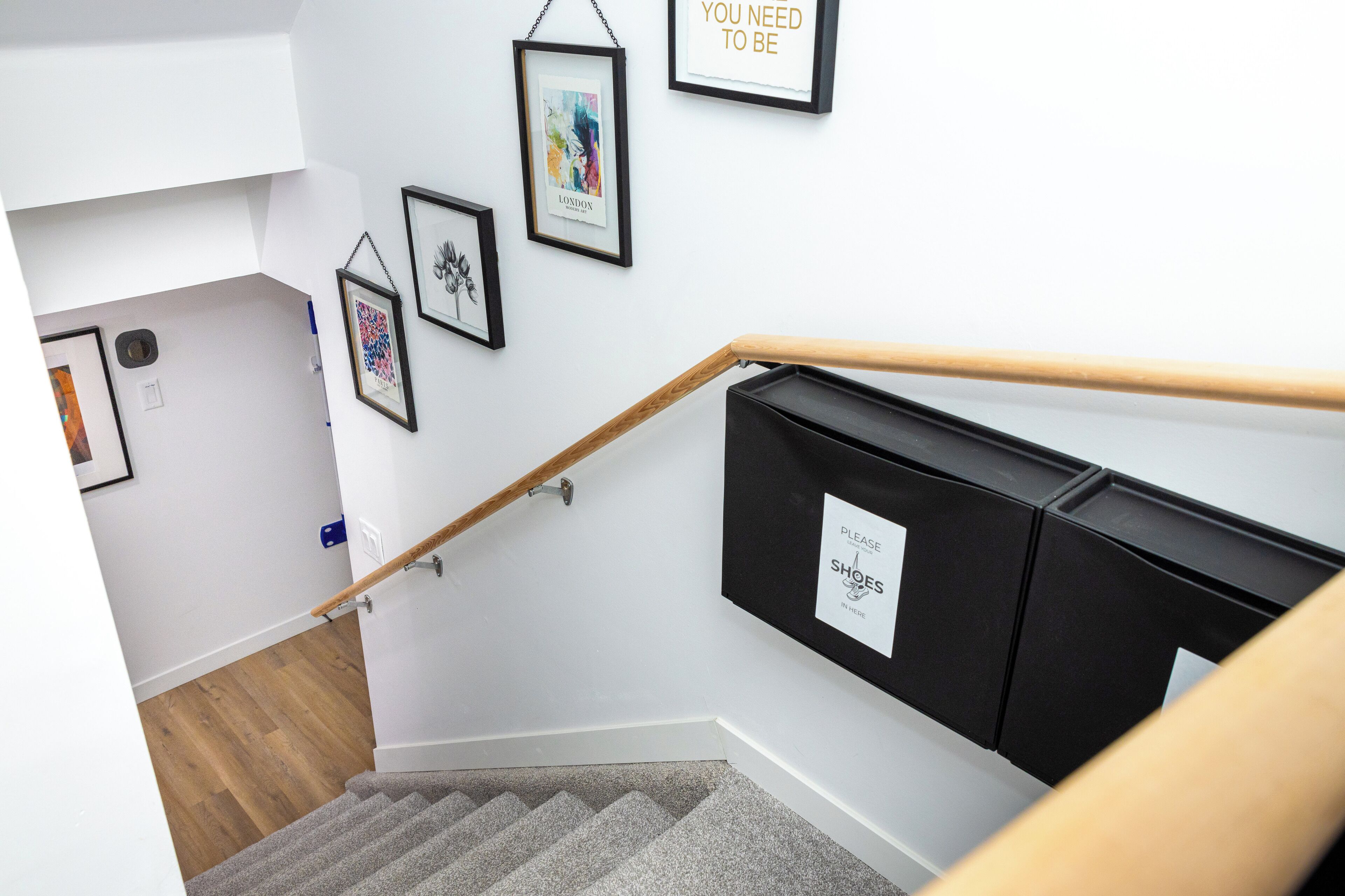 Un escalier avec une rampe en bois, un meuble noir pour ranger les chaussures et des tableaux encadrés accrochés au mur.
