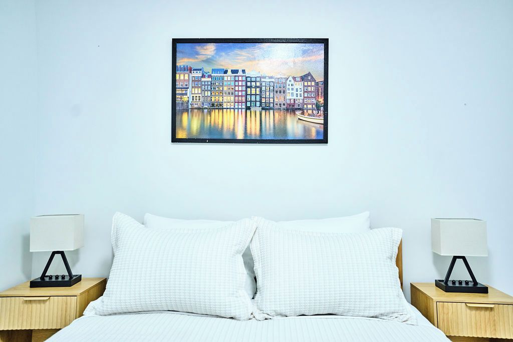 Une chambre d’hôtel avec un lit, deux tables de chevet avec des lampes et un tableau représentant un paysage urbain, encadré et accroché au mur.