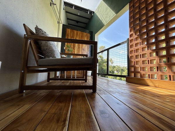 Eine Holzbank mit Kissen auf einem Balkon mit Holzboden, Blick ins Grüne und eine Backsteinwand mit Gittermuster.
