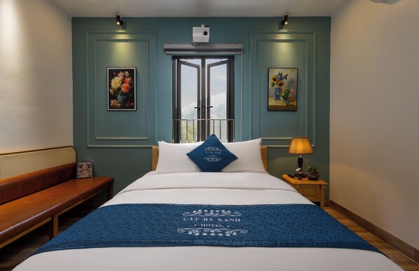 A hotel room with a bed, a bench, a nightstand with a lamp, and two framed floral paintings on the walls. - Cat Ba Xanh Spring Garden Hotel  (Hai Phong)